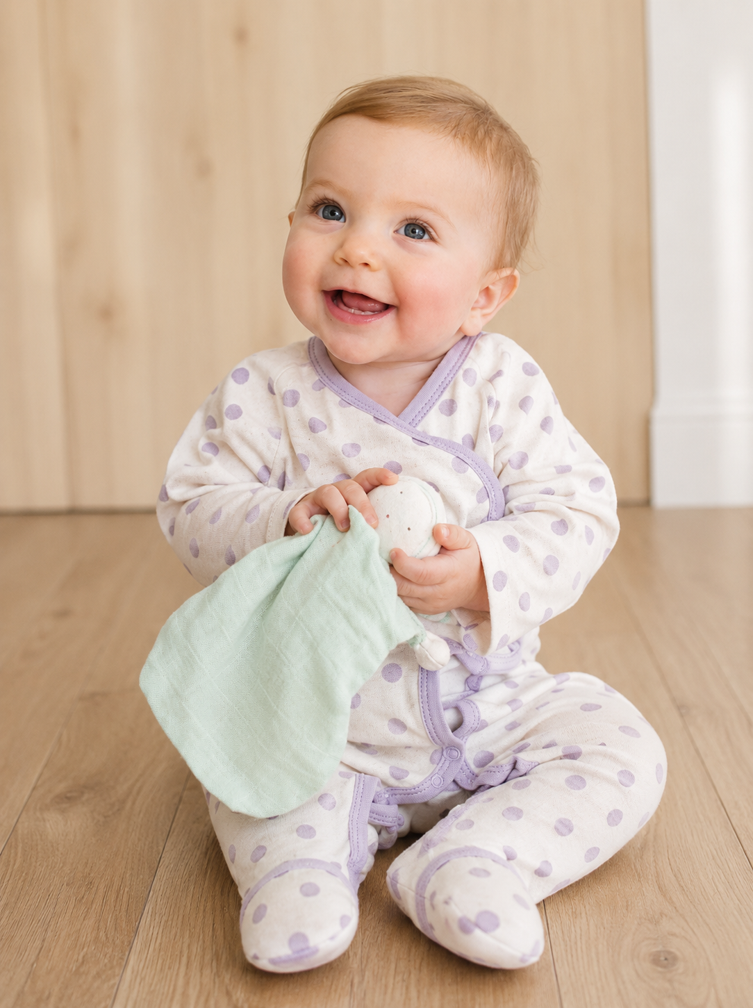 Smiling baby wearing an organic baby footie in GOTS-certified organic Egyptian cotton, with a lavender polka dot print and side-snap wrap front