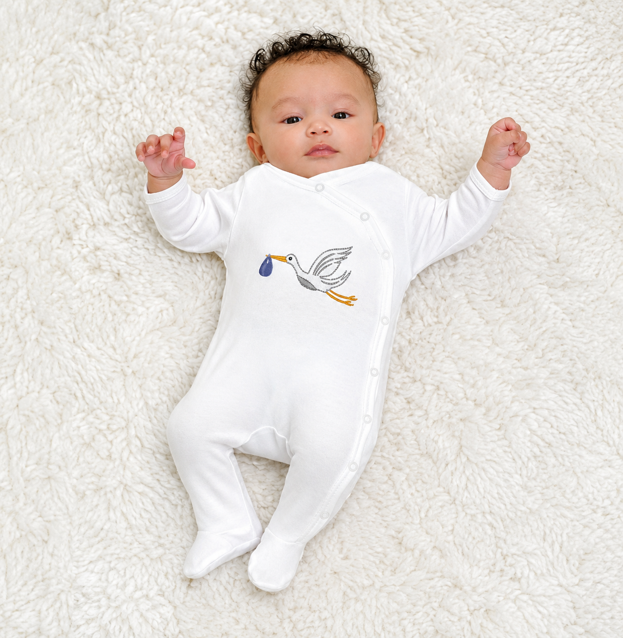 Newborn baby lying down wearing an organic newborn sleepsuit with stork embroidery and side snap closure